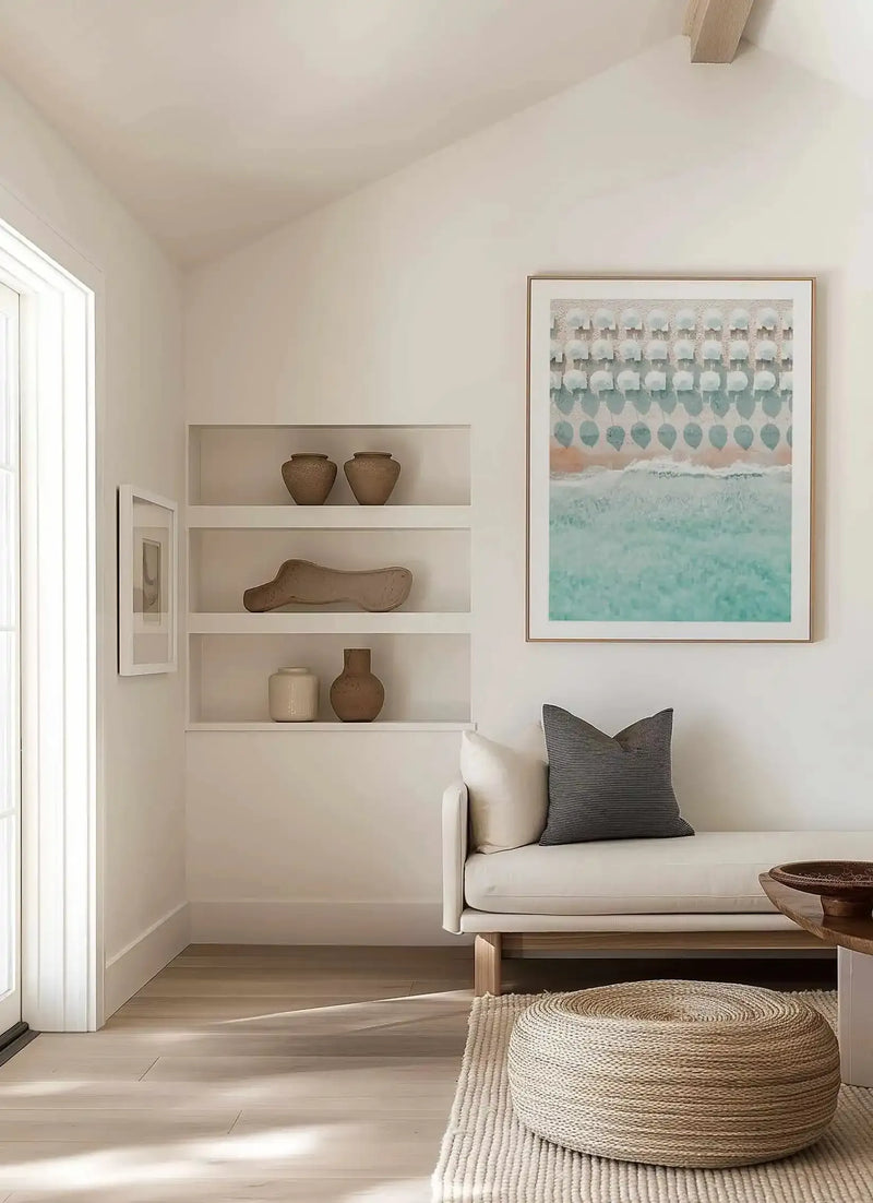 Lifestyle shot of a minimalist living room featuring a large framed art print of an aerial beach scene with white umbrellas. The print depicts rows of white umbrellas on a light sand beach, with gentle turquoise waves breaking on the shore, displayed in a natural wood frame with a white border. The room includes a cream sofa with a dark grey striped cushion, a woven jute pouf on a textured rug, and built-in shelves with decorative pottery.
