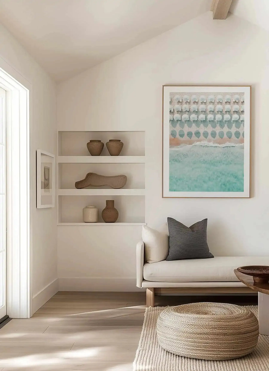 Lifestyle shot of a minimalist living room featuring a large framed art print of an aerial beach scene with white umbrellas. The print depicts rows of white umbrellas on a light sand beach, with gentle turquoise waves breaking on the shore, displayed in a natural wood frame with a white border. The room includes a cream sofa with a dark grey striped cushion, a woven jute pouf on a textured rug, and built-in shelves with decorative pottery.