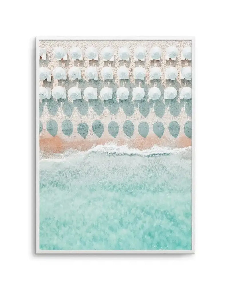 Framed aerial photograph of a beach scene with white frame and white border. The composition features rows of white beach umbrellas casting long, light blue shadows on the pale sand, with gentle turquoise waves breaking on the shore.