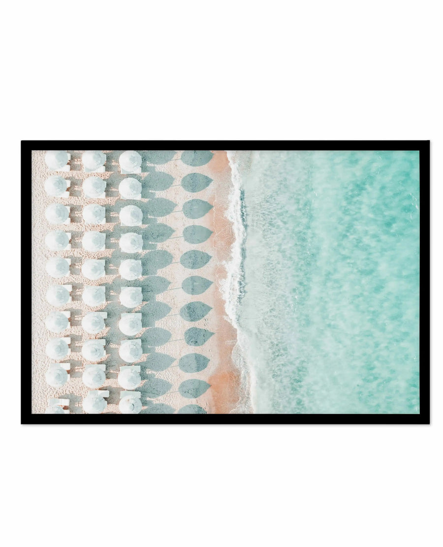 Aerial view art print featuring rows of white umbrellas casting teal and gray shadows on sandy beach, with turquoise ocean water and white foam at the shoreline. Photograph-style image in black frame with white border, captured from directly above showing organized beach setup with rippled water texture and natural coastal palette of cream, tan, and aquamarine tones.