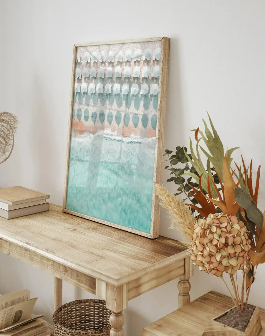 Lifestyle shot of a natural wood console table with a framed art print leaning against a white wall. The art print, titled "White Umbrellas", depicts an aerial view of a beach with rows of white umbrellas on light tan sand, leading to turquoise ocean waves. The print is in a natural wood frame with a white border. To the left of the print are three stacked books, and to the right is a vase with dried pampas grass and other dried foliage in shades of orange, green, and beige.