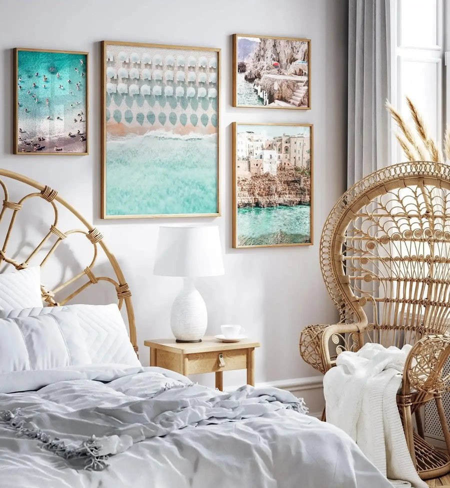 Lifestyle shot of a bedroom featuring a framed art print of white umbrellas on a beach, displayed above a wooden bedside table. The print is a top-down photographic view of a beach with turquoise water and white sand, featuring rows of white umbrellas and people swimming and sunbathing. The room includes a rattan headboard, a white bedspread, a white table lamp, and a large rattan peacock chair.