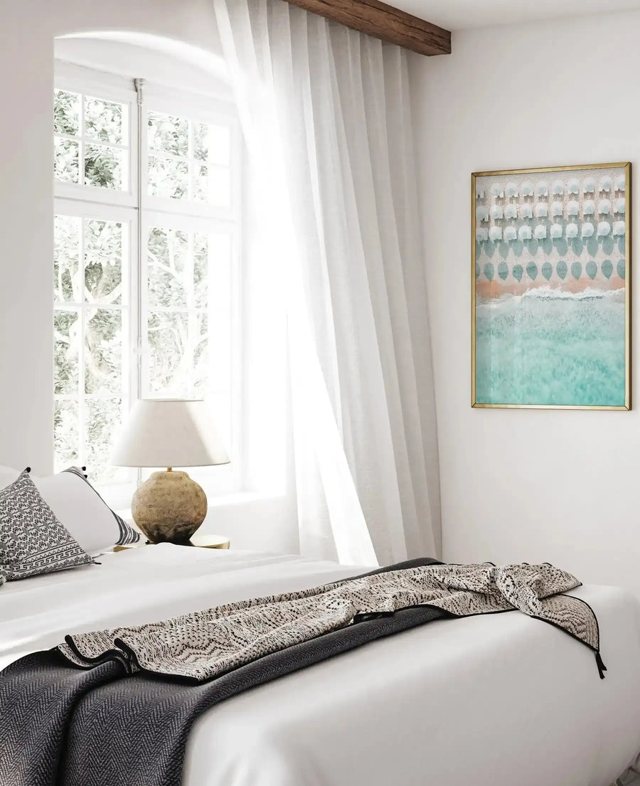 White Umbrellas art print displayed in a gold frame on a white bedroom wall above a bed with charcoal and cream textiles. The watercolor artwork features a repetitive pattern of white umbrellas arranged in rows against soft blue-green and peachy-beige horizontal bands, creating a serene, minimalist composition. The framed print hangs beside a bright window with flowing cream curtains and wooden beam, in a calm, Scandinavian-inspired bedroom setting.