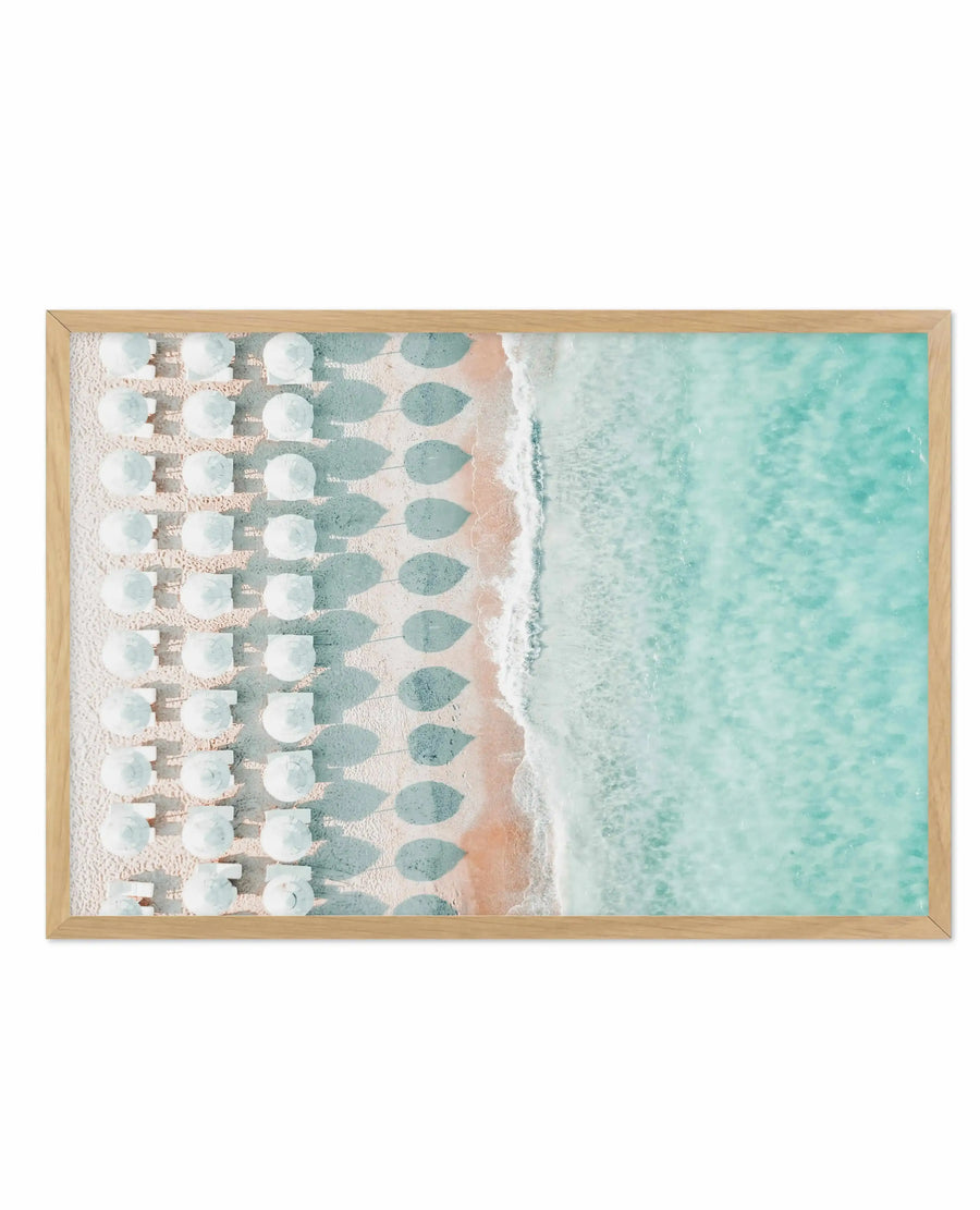 Aerial photograph framed in natural wood showing a beach scene with rows of white and teal umbrellas lined up on pale sand next to turquoise shallow water. The composition is divided vertically with the organized umbrella pattern on the left and open water on the right. The image has a soft, muted color palette of cream, dusty teal, and peachy-beige tones with a watercolor-like quality. The natural wood frame has a warm honey finish.