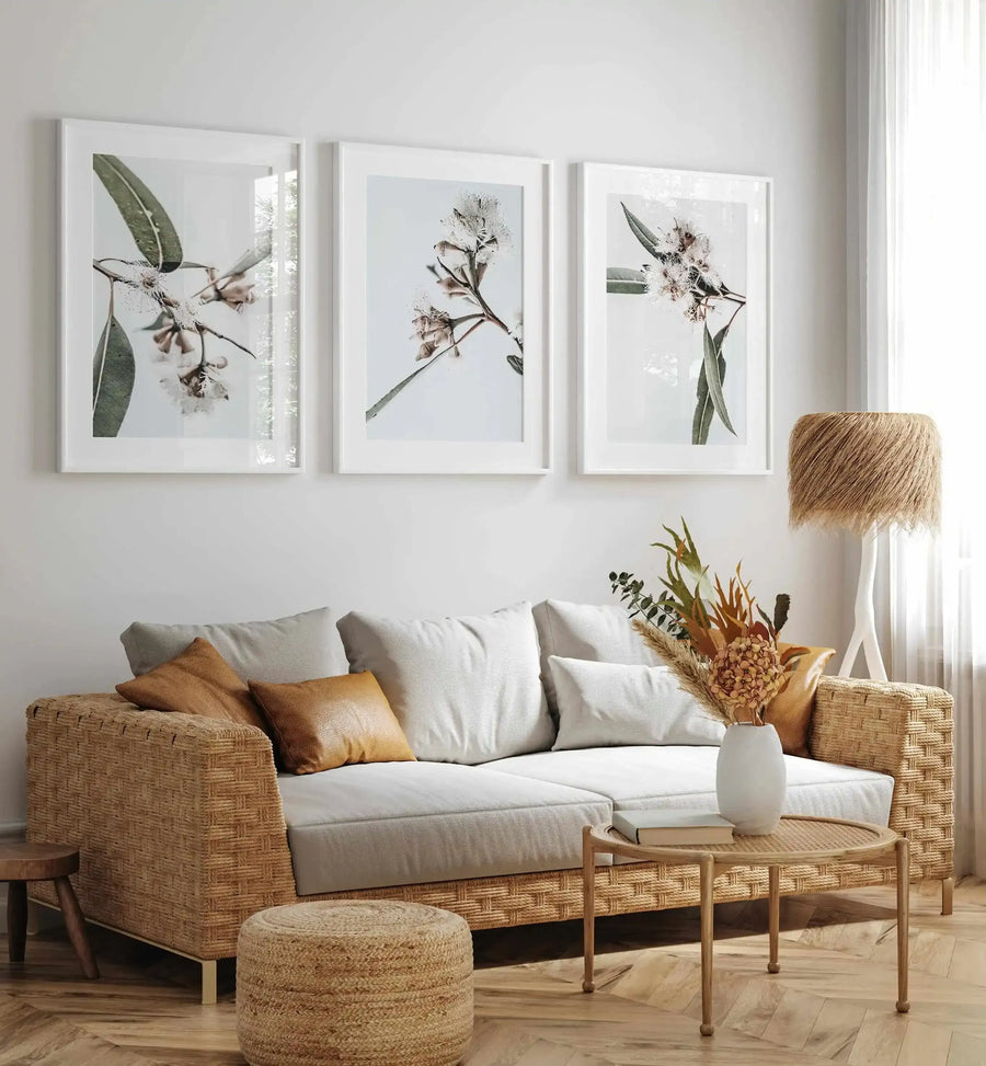 Lifestyle shot of three framed art prints displayed above a woven rattan sofa in a bright, minimalist living room. Each print features a close-up botanical photograph of eucalyptus branches with delicate white flowers and long, slender green leaves against a soft white background, with white frames and white borders. The room includes a light grey sofa with cream and tan cushions, a round rattan coffee table with dried floral arrangement, a woven ottoman, and a tall floor lamp with a textured, shaggy shade.