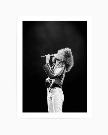 Framed black and white photograph of Whitney Houston singing into a microphone on stage, with a dark background. She is captured mid-performance, looking upwards with her eyes closed, wearing a long-sleeved top and light-coloured jeans, with her curly hair flowing. The print is displayed with a white border.