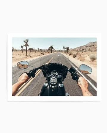 "Art print featuring a first-person perspective photograph of a person riding a motorcycle down a desert highway. The road stretches into the distance, flanked by sandy terrain and numerous Joshua trees under a clear, bright blue sky. The rider's hands are visible on the handlebars, with the motorcycle's dashboard and windshield in the foreground. The rearview mirrors reflect the rocky desert landscape."