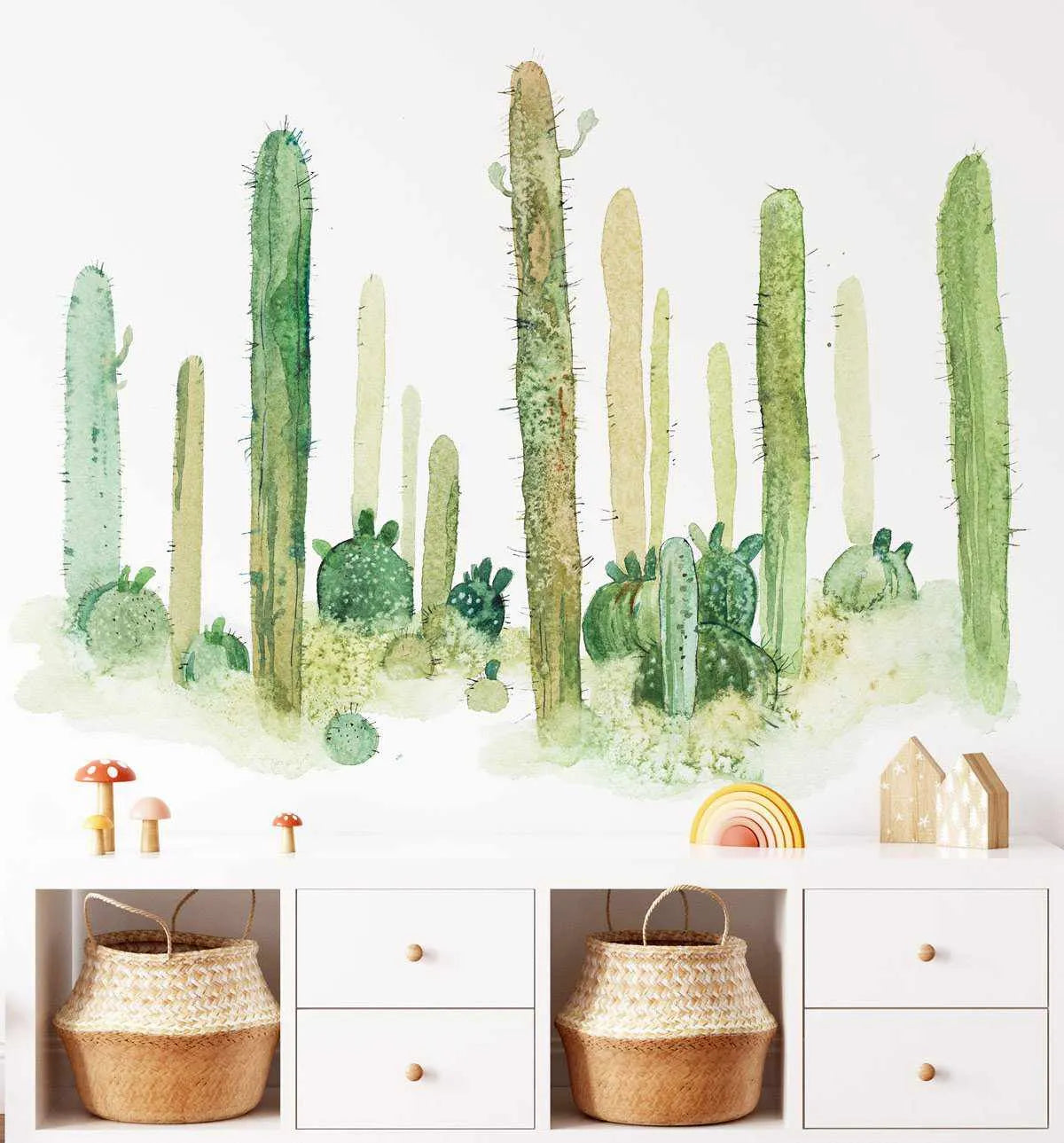 Kids' Room:
"Wild Cactus Wallpaper Mural displayed on a white wall above a low white storage unit in a child's room. The peel and stick wallpaper features a desert landscape with various hand-painted cacti in shades of green and beige, with visible watercolour brushstrokes and textured ground. The storage unit holds two woven baskets and is decorated with wooden mushroom figures, a rainbow toy, and wooden house ornaments."