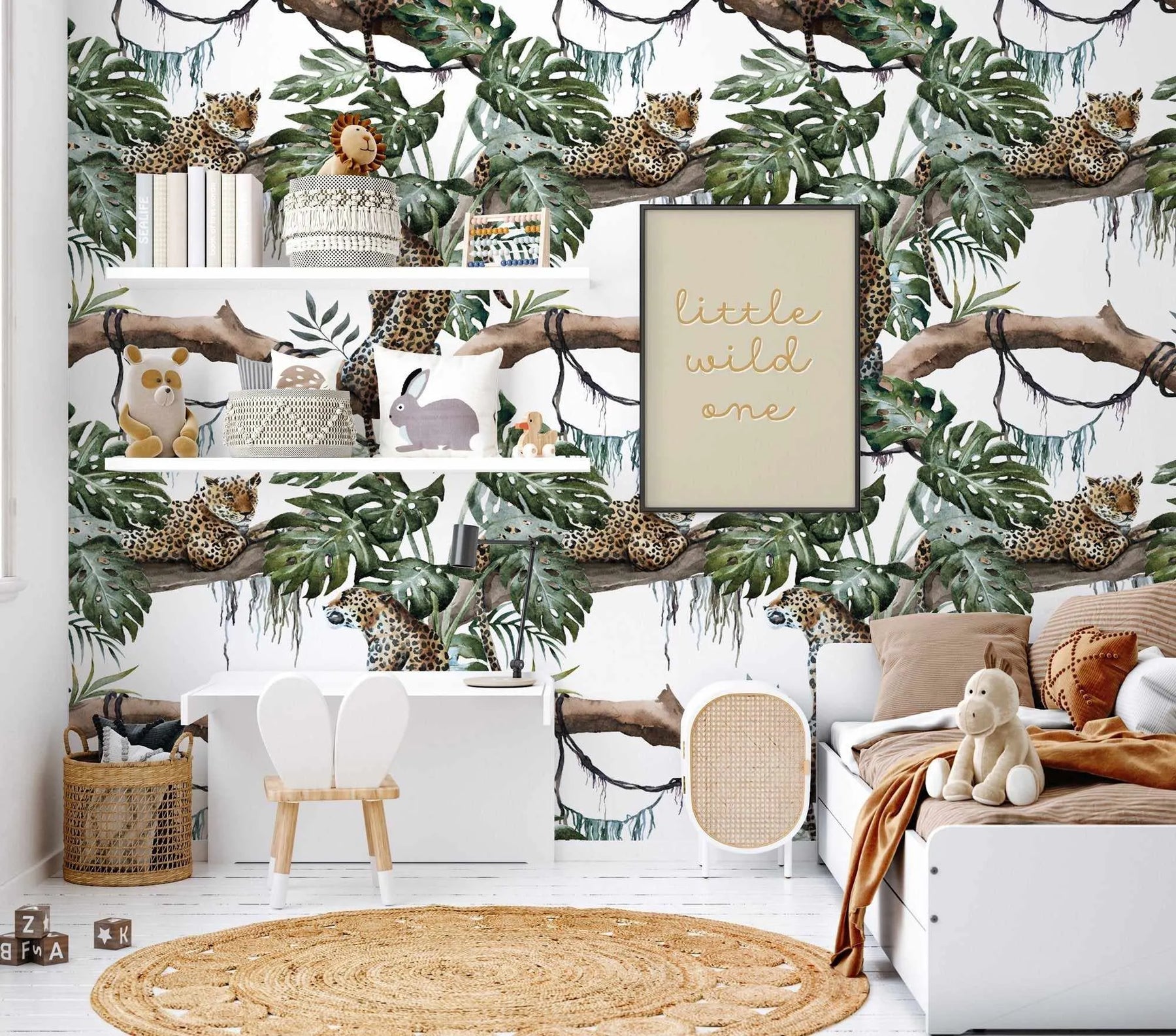 Lifestyle shot featuring a child's bedroom with a jungle-themed wallpaper. The wallpaper features a repeating pattern of hand-painted leopards resting on tree branches amidst large green monstera leaves and hanging vines on a white background. The room includes a white desk with two white bunny-ear chairs, a white bed with a brown duvet and pillows, a framed print with the words "little wild one" in gold script, and various toys and baskets.