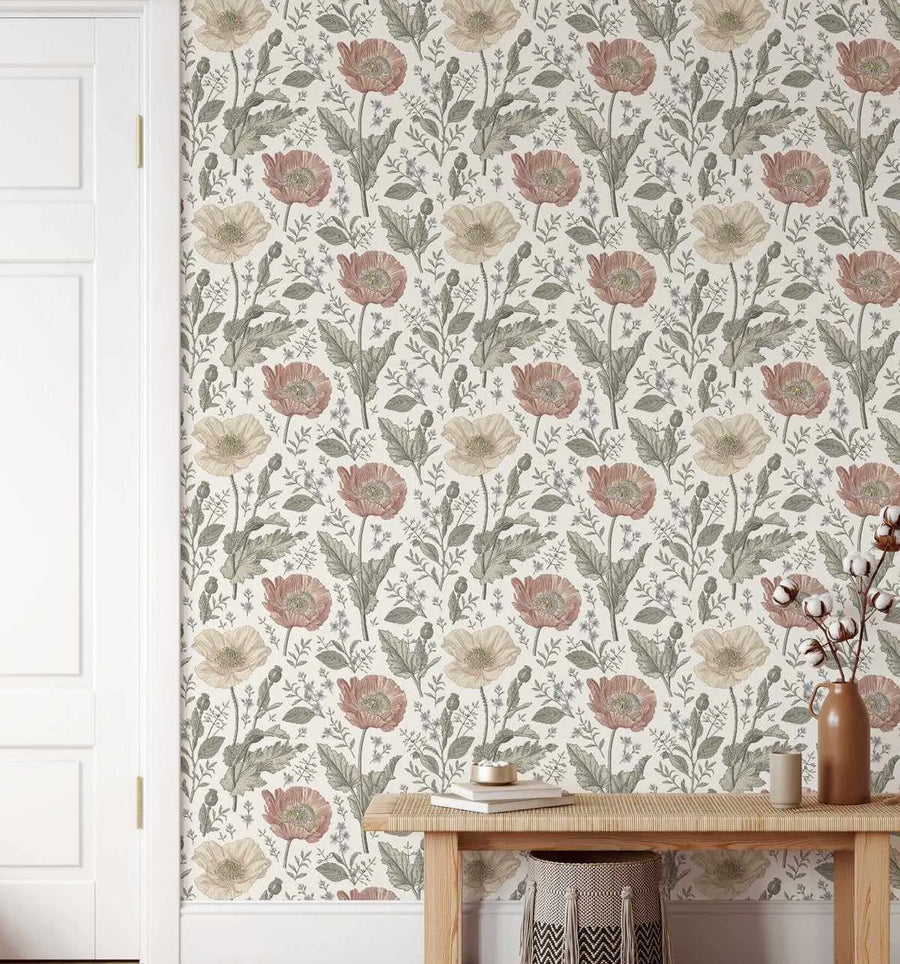 Wallpaper featuring a repeating pattern of hand-drawn poppy flowers in muted shades of dusty rose, pale cream, and soft beige, with sage green leaves and delicate blue sprigs on a light cream background. The botanical design has a detailed, illustrative quality. The wallpaper is shown on a wall next to a white door and above a light wood console table with a woven basket underneath.