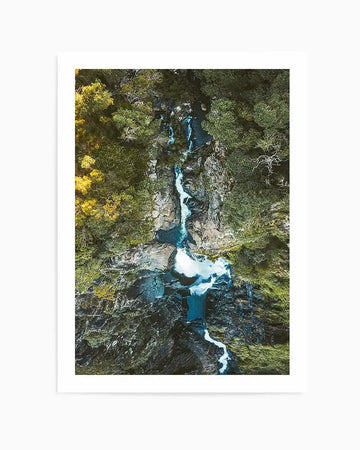 "Aerial photography art print of a winding waterfall cascading through a lush green and yellow forest, printed on fine art paper and displayed with a white border. The waterfall flows over dark, rocky terrain, creating white foamy pools at various points before continuing its journey downstream."
