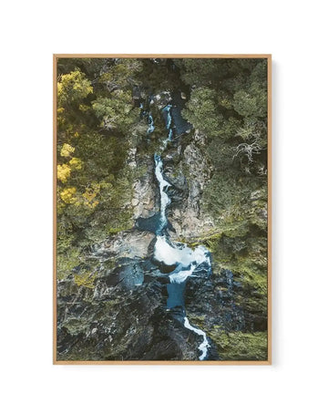 Aerial view photograph of Windin Falls near Cairns displayed in a natural wood frame. The image shows a dramatic narrow gorge carved through grey rock formations, with a turquoise-blue river winding through the center. Lush green forest canopy surrounds the gorge on both sides, with lighter yellow-green foliage visible on the left. White water rapids are visible along the river's path. The photograph is a vertical composition printed on canvas with a natural wood-toned frame.