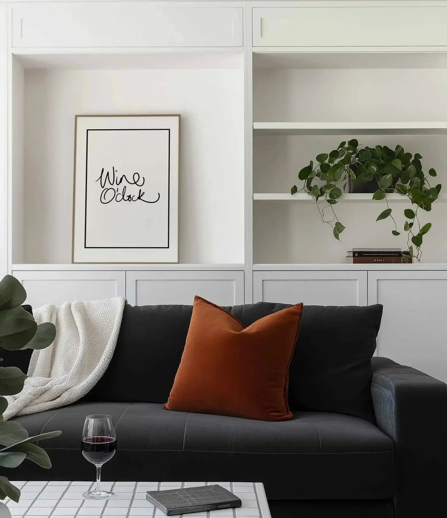 Wine O'Clock art print displayed in a natural wood frame with white border, hung in a white built-in shelving unit above a black sofa. The print features handwritten-style black text reading "Wine O'Clock" on a cream background. Room styling includes an orange rust velvet cushion, cream knit throw, trailing potted plant on upper shelf, and a wine glass on the table below.