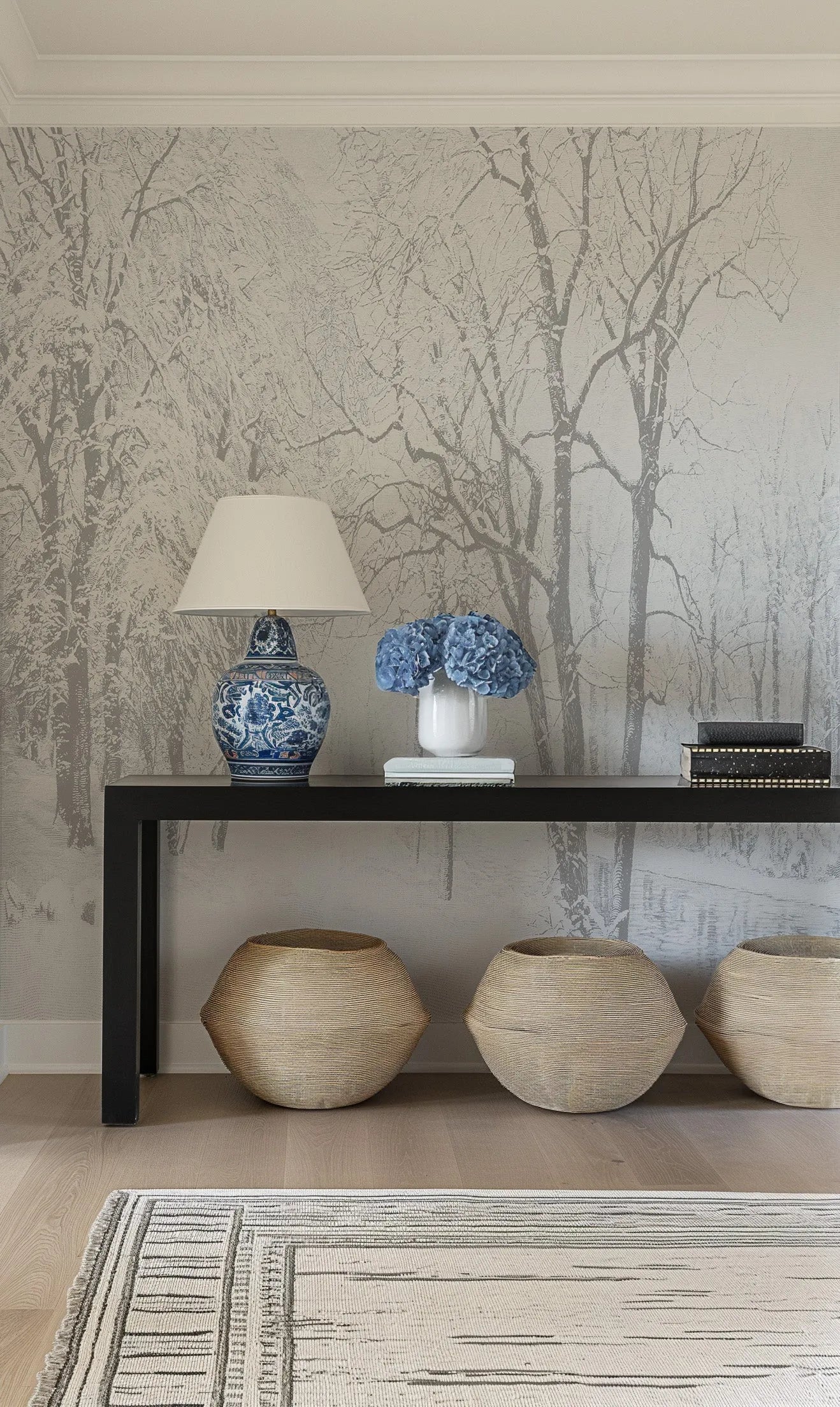 Hallway:
"Winter Wonderland Wallpaper Mural displayed in a hallway with a black console table, a blue and white patterned lamp, and three woven baskets. The wallpaper features a repeating pattern of a snowy forest with bare trees in shades of grey and white, creating a serene and wintry landscape. The console table holds a white vase with blue hydrangeas and a stack of books, while a striped rug is partially visible on the light wood floor."