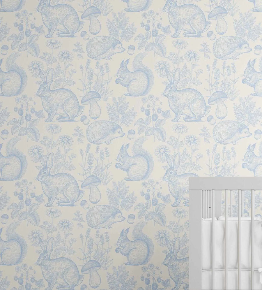 Woodland Folk Wallpaper in Sky Blue featuring a repeating pattern of forest animals and botanicals rendered in soft blue line drawings on a cream background. The design includes rabbits, squirrels, hedgehogs, and mushrooms interspersed with delicate wildflowers, ferns, and botanical elements. The illustration style appears hand-drawn with fine linear detailing. A white painted crib is visible in the lower right corner, showing how the wallpaper coordinates with nursery furnishings.