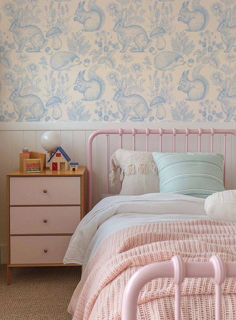 "A child's bedroom featuring wallpaper with a repeating pattern of woodland animals and plants in sky blue on a cream background. The design includes rabbits, squirrels, hedgehogs, mushrooms, daisies, and lavender. The wallpaper is displayed above white wainscoting and a pink metal bed frame with white bedding, a cream textured throw, and a light blue striped pillow. A light wood bedside table with three blush pink drawers and brass knobs holds a white globe lamp and wooden toys."
