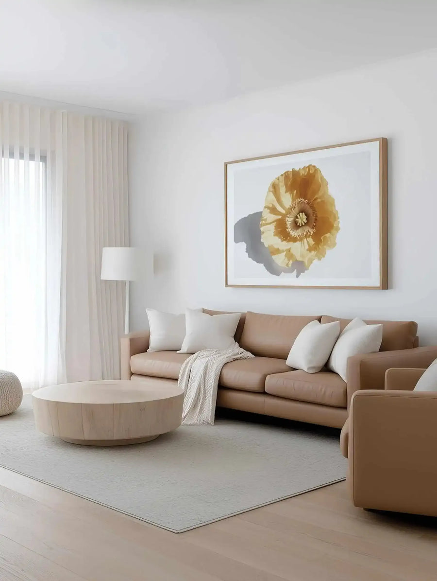 Yellow Poppy Art Print in natural wood frame displayed on white wall above tan leather sofa in minimalist living room. The artwork features a large golden-yellow poppy flower rendered in watercolor style with delicate petals and dark center, set against a white background with soft gray shadow. The print has a white border and is framed in light natural wood. The room includes cream vertical blinds, round cream coffee table, white floor lamp, beige area rug, and white throw pillows.