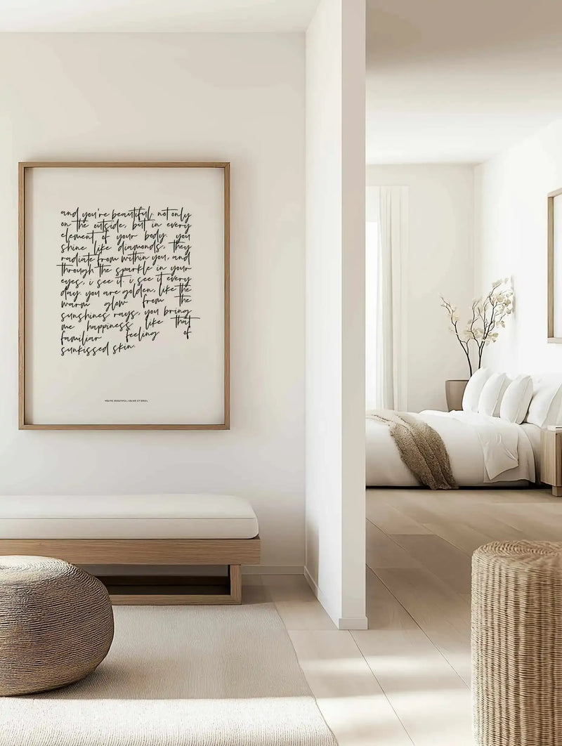 Framed art print displaying handwritten black script text on cream background, mounted in natural wood frame with white border. Text reads an inspirational poem about inner beauty and radiance. Print displayed on white bedroom wall above a wooden bench with white cushion. Adjacent room shows white bedding, decorative pillows, dried branches in vase, and woven basket accents. Serene, minimalist aesthetic with warm neutral tones throughout the space.