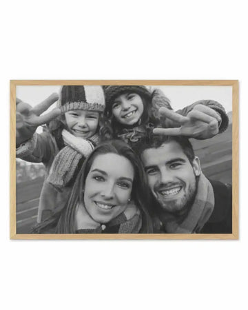 Framed black and white photograph of a family of four smiling and posing for a selfie, with a natural oak frame and white border. The composition features two adults and two children in winter clothing, with the children making peace signs with their hands.