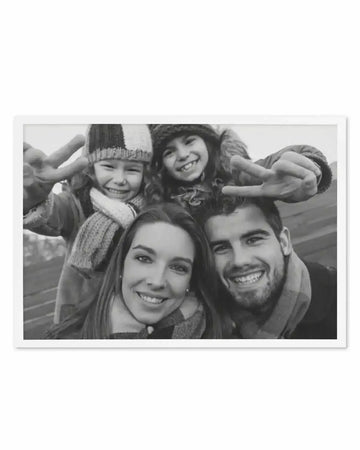 Framed black and white photograph of a family of four taking a selfie, displayed in a white frame with a white border. The composition features two adults and two children smiling at the camera, with the children holding up peace signs.