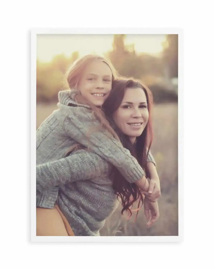 Framed photography art print featuring a mother giving her daughter a piggyback ride in a field at sunset, with a white frame and white border. The mother, with long dark hair, smiles at the viewer while her daughter, with long blonde hair, rests her head on her mother's shoulder and smiles. Both wear grey knit sweaters.
