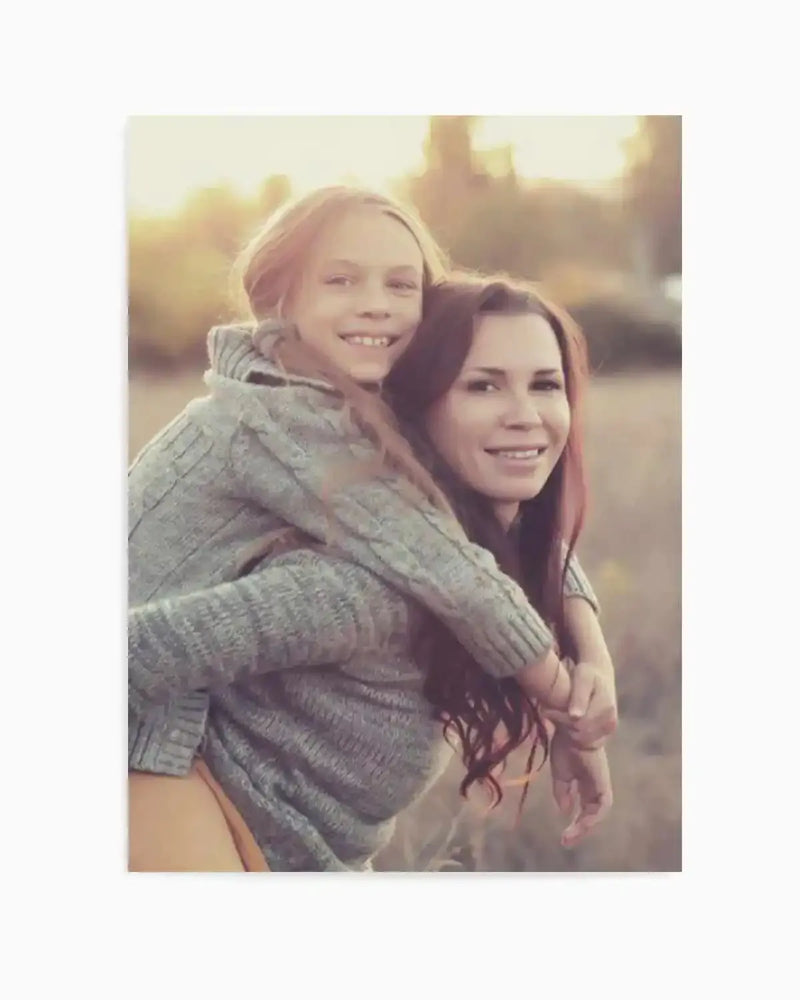 Framed photography art print of a young girl with long blonde hair giving a piggyback ride to a woman with long brown hair, both smiling and wearing grey knitted sweaters, against a soft-focus outdoor background with warm, golden light, displayed in a white frame with a white border.