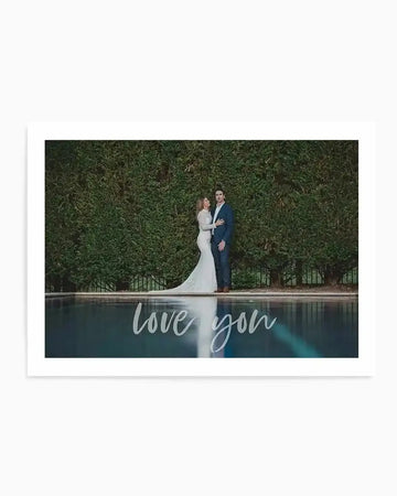 Personalized art print featuring a wedding photo of a couple standing by a poolside with a tall green hedge wall backdrop. The bride wears a white mermaid gown and the groom wears a dark navy suit. Script text reading "love you" overlays the lower portion of the image in white handwriting. The print is mounted on a white background with a white border, displayed as a horizontal landscape-oriented artwork.
