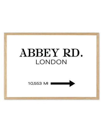 Art print featuring the words "ABBEY RD. LONDON" in large black serif font, with "10,553 MI" and a black arrow pointing right below, all on a white background, displayed in a natural oak frame with white border.