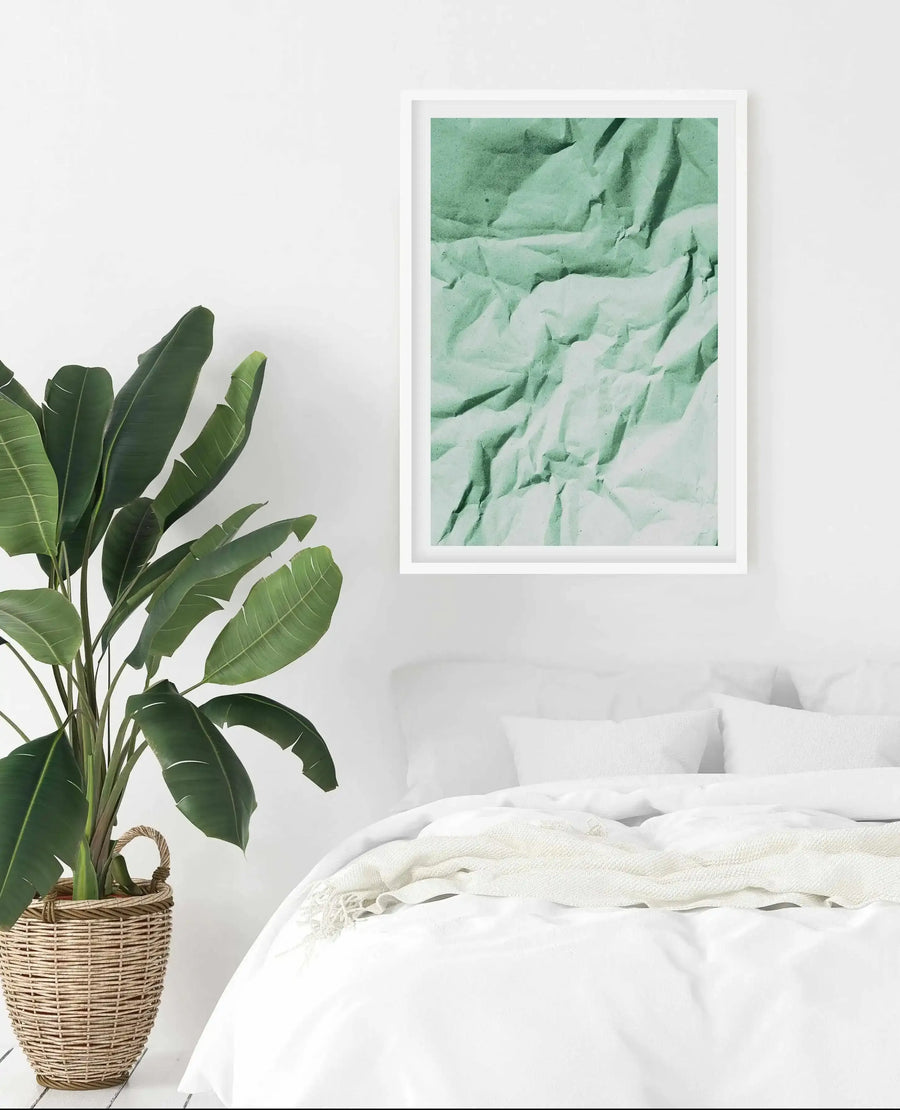 Large framed art print displayed above a bed in a minimalist bedroom. The print features a crumpled sheet of paper in a muted sage green with deep shadows and highlights creating a textured, abstract composition, with a white frame and white border. The room includes a large green plant in a woven basket and a bed with white linen and a cream throw.
