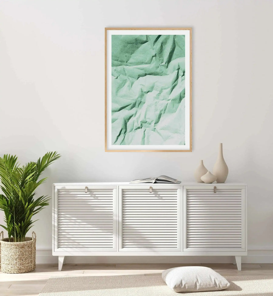 Lifestyle shot of a minimalist living room with a framed art print featuring crumpled paper texture in a muted sage green, displayed above a white slatted console table. The room includes a large potted palm plant in a woven basket, cream ceramic vases, and a striped cushion on a light rug.