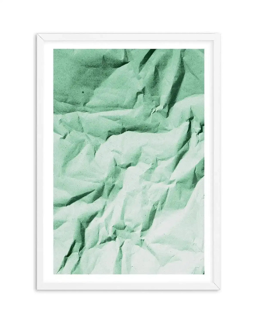 Abstract art print featuring a crumpled paper texture in a muted sage green, with darker shadows and lighter highlights creating a dynamic, textured surface. The print is displayed in a white frame with a white border.