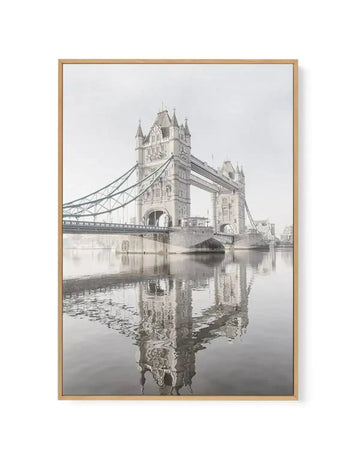 Framed black and white photograph of the Tower Bridge in London, printed on canvas and displayed in a natural oak frame. The composition features the iconic bridge with its two prominent towers and suspension cables, reflected in the calm water of the River Thames below, under a bright, overcast sky.