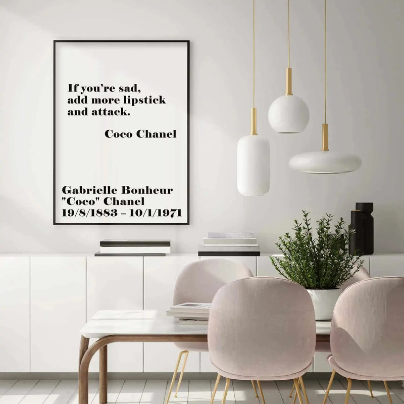 Framed art print with black frame displaying a Coco Chanel quote in bold serif typography on white background. Quote reads "If you're sad, add more lipstick and attack." attributed to Gabrielle Bonheur "Coco" Chanel with birth and death dates 19/8/1883 – 10/1/1971. Print shown in minimalist dining room setting with white pendant lights, natural wood table, blush pink chairs, potted green plant, and stacked books on white storage unit.