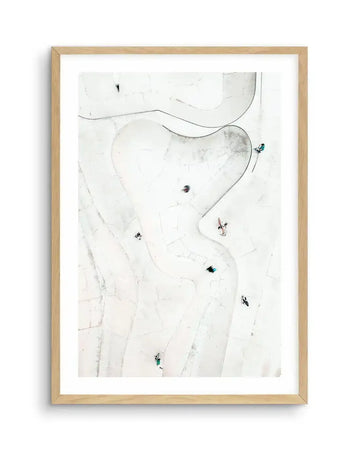 Framed aerial photography art print of a white concrete skate park with several people in motion, displayed in a natural oak frame with white border. The composition features a winding, organic layout of ramps and bowls with subtle cracks and shadows on the bright white surface.