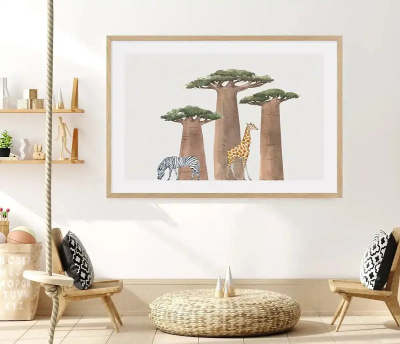 Lifestyle shot of a child's room featuring a large framed art print of an African landscape displayed on a white wall above a low woven coffee table. The print features four baobab trees with large trunks and flat green canopies, with a zebra grazing on the left and a giraffe standing on the right. The art print has a natural wood frame and a white border. The room includes two small wooden chairs with black and white patterned cushions, a rope swing, and shelves with various decorative items.