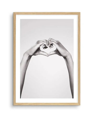 Framed black and white photograph of two hands forming a heart shape against a plain light grey background, with natural wood frame and white border. The hands are positioned centrally, fingers touching to create the top curves of the heart, and wrists meeting at the bottom point.
