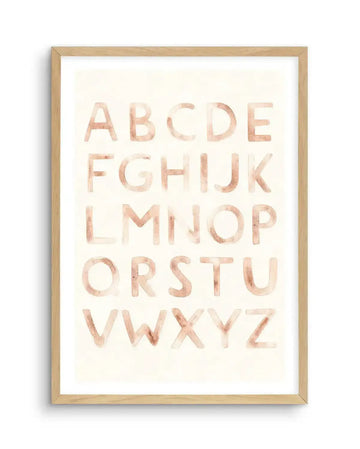 Watercolour art print featuring the complete alphabet in uppercase letters, painted in a soft, muted blush pink with visible brushstroke texture on a warm cream background, displayed in a natural oak frame with white border.
