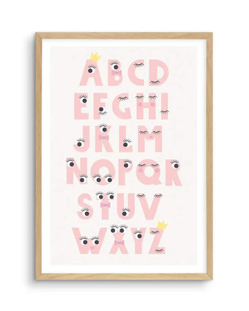Art print featuring the alphabet in large, playful pink capital letters, each adorned with cartoon eyes and long eyelashes, some with small black pupils and others with closed eyelids. The letters also include small details like a yellow crown on 'A' and 'Y', and pink bows on 'B', 'K', 'S', and 'X'. The print is displayed in a natural oak frame with a white border.