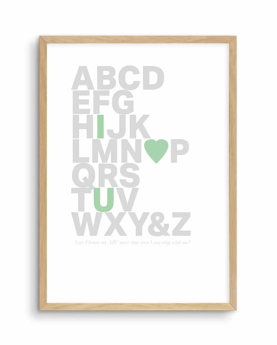 Framed art print featuring the complete alphabet in large light gray letters arranged vertically, with select letters highlighted in soft sage green: the letter I, a heart symbol replacing O, and the letter U. At the bottom, small gray italicized text reads "Now I know my ABC, next time won't you sing with me?" The artwork is displayed in a natural wooden frame with a white background.