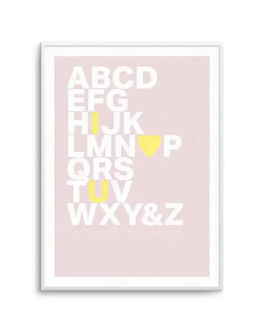 Framed art print featuring the full alphabet arranged vertically on a soft dusty rose pink background. White capital letters spell out ABCCD EEFG HIJKL LMNO PQRS TUVW XY&Z with yellow heart shapes and yellow rectangular blocks replacing certain letters. Small white text appears at the bottom. The print is displayed in a white frame with white border.