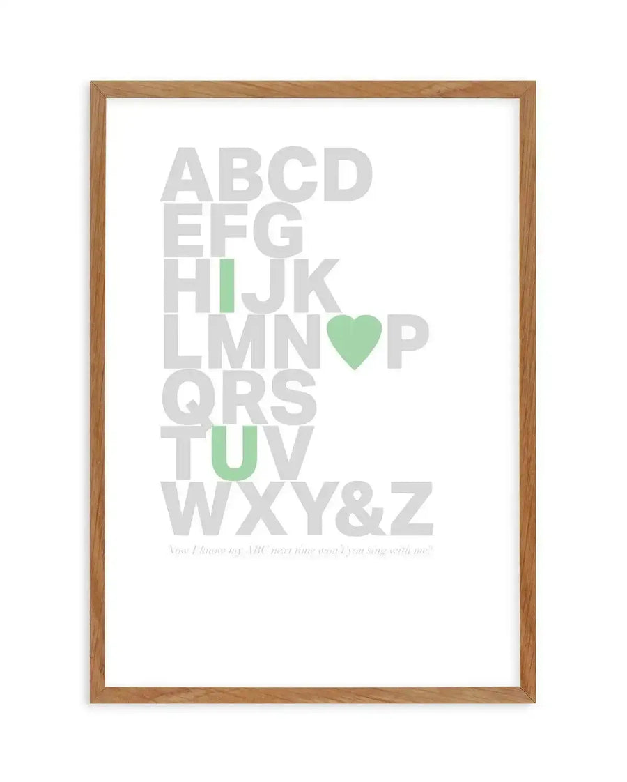 Framed art print featuring the alphabet in large light gray letters arranged vertically. The letters I, O, and U are highlighted in soft sage green, as is a green heart symbol replacing the O in "LMNO." Below the alphabet, small italicized text reads "You I know my ABC next time won't I you sing with me?" The print is displayed in a natural wood frame with a white background.