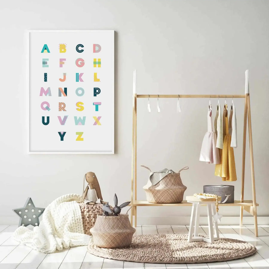 Lifestyle shot of a child's room featuring a framed alphabet art print displayed on a light grey wall. The print features uppercase letters A-Z in a grid, each letter a different colour and pattern, including teal, yellow, pink, and black with stripes, dots, and geometric shapes. The room includes a light wood clothing rack with children's dresses, woven baskets with toys and blankets, a white stool, and a round braided rug on a white wooden floor.
