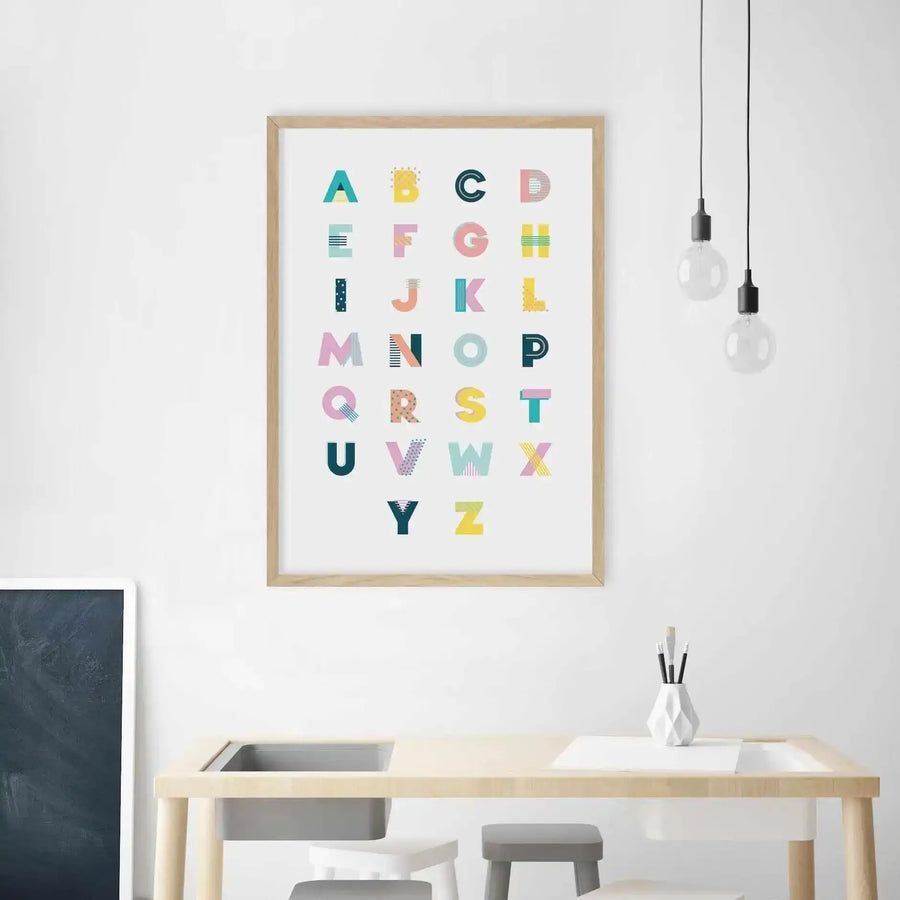 Kids Room:
"Framed alphabet art print displayed above a light wood children's activity table in a minimalist room. The print features the English alphabet from A to Z, with each letter in a different bright colour and pattern, including teal, yellow, pink, and blue. The room includes a dark grey chalkboard, white and grey stools, and a white geometric vase with paintbrushes."