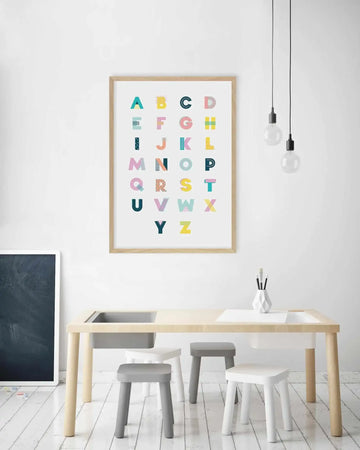 Kids Room:
"Framed art print featuring a colourful alphabet displayed above a wooden play table in a bright kids' room. The print showcases each letter of the alphabet in a different pastel colour and geometric pattern, with a natural wood frame and white border. The room includes a black chalkboard, a white geometric vase with pencils, and four small stools in white and grey."