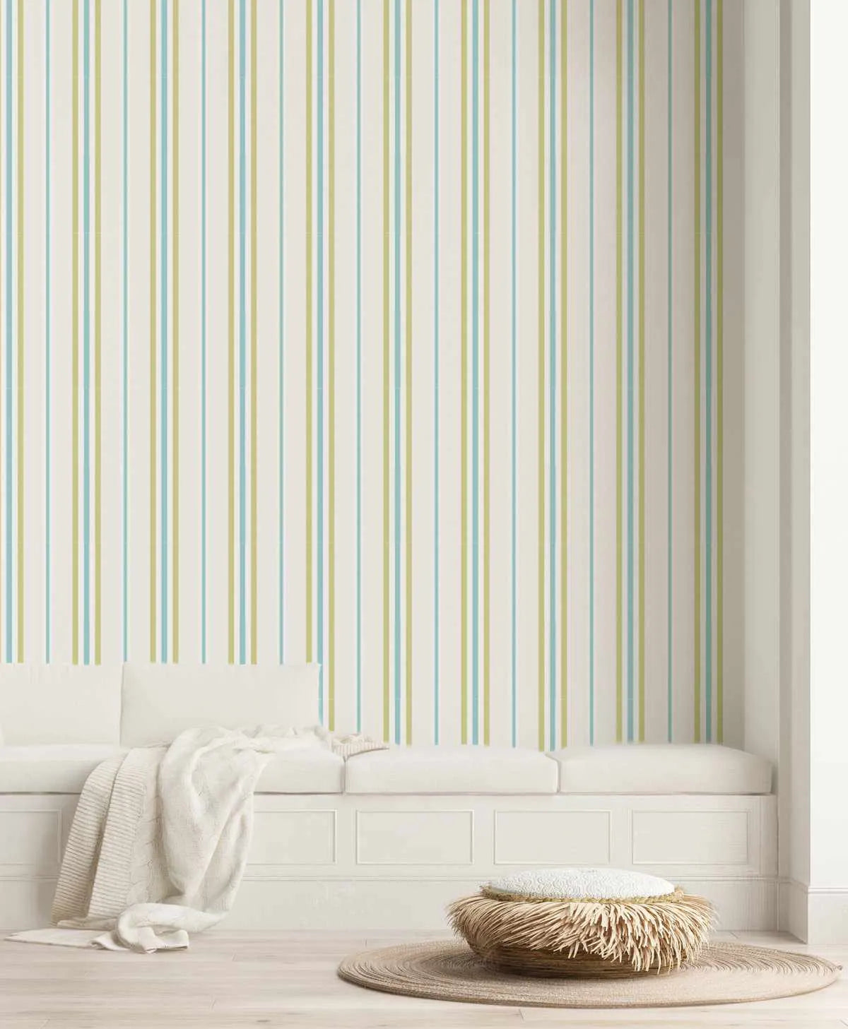 Lifestyle shot of a room with Amalfi Stripe Wallpaper. The wallpaper features vertical stripes in alternating pale blue and muted sage green on a cream background. The room includes a white built-in bench with cushions and a cream knitted throw, a woven round rug, and a textured ottoman.