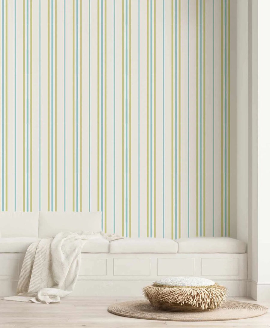 Lifestyle shot of a room with Amalfi Stripe Wallpaper. The wallpaper features vertical stripes in alternating pale blue and muted sage green on a cream background. The room includes a white built-in bench with cushions and a cream knitted throw, a woven round rug, and a textured ottoman.