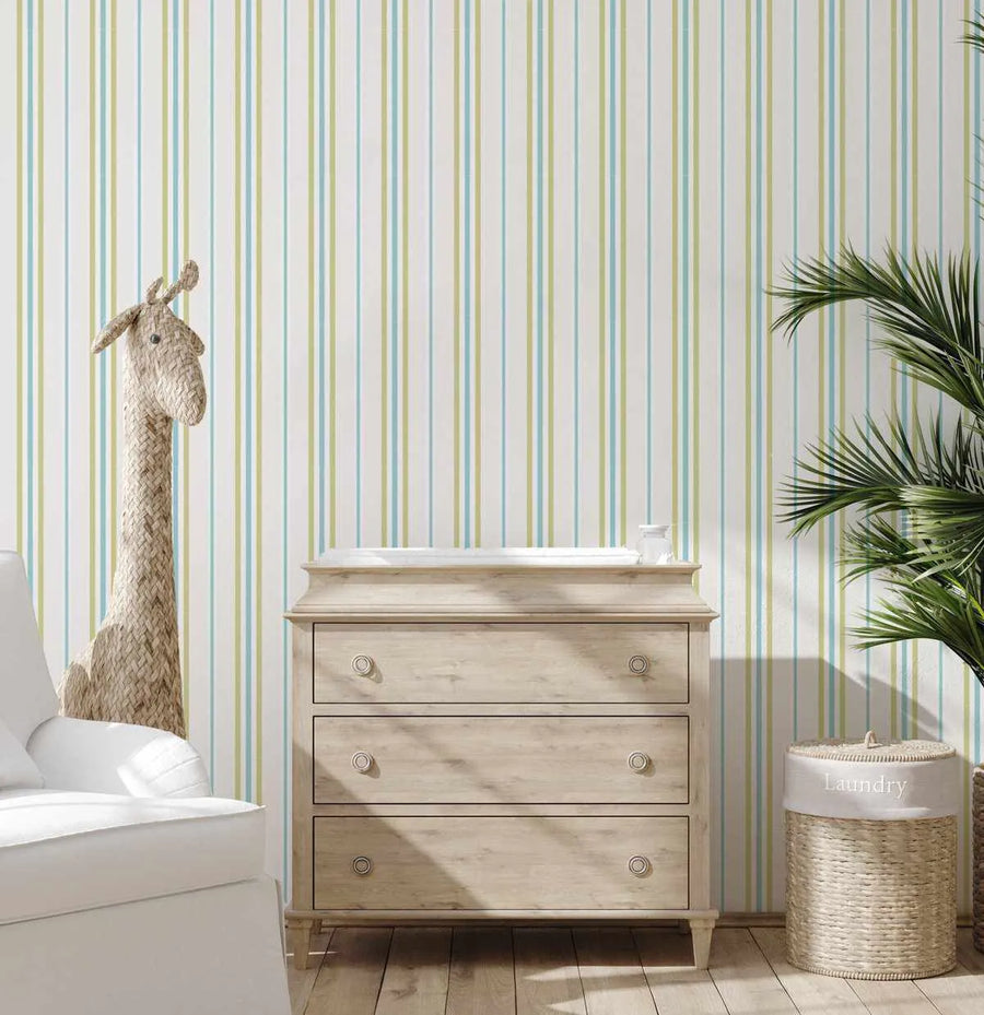Amalfi Stripe Wallpaper displayed on a bathroom wall featuring vertical stripes in soft blue-green, pale yellow, and cream tones with consistent narrow line spacing. The wallpaper is shown in a styled bathroom setting with a whitewashed wooden three-drawer dresser, woven giraffe decoration, potted palm plant, and woven laundry basket, demonstrating the wallpaper's coastal, relaxed aesthetic against pale wood flooring.