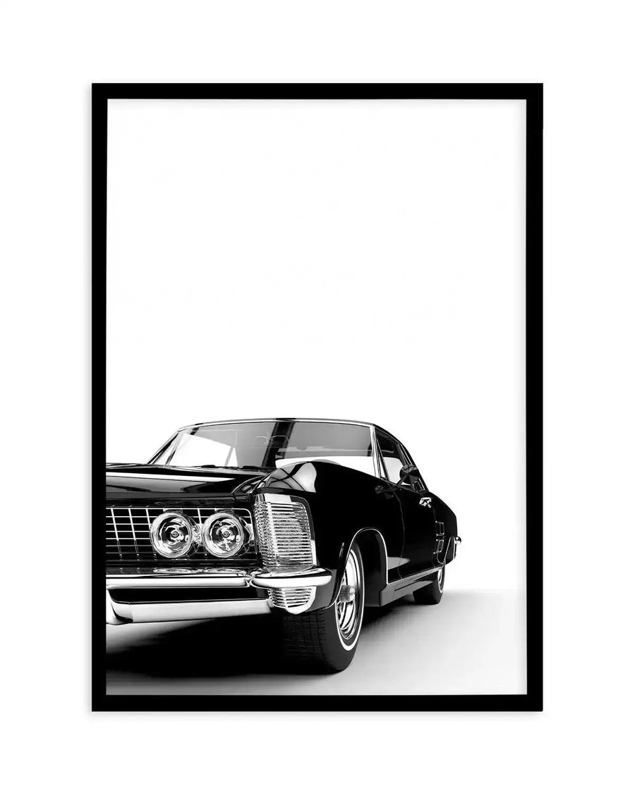Black and white photograph of a vintage American muscle car displayed in a black frame with white border. The car, positioned at a three-quarter angle facing left, features dual round headlights, a horizontal grille, chrome bumper and trim details, and an open driver's side door. The vehicle is rendered in high-contrast black and white photography against a plain white background. The frame is simple and modern with clean black edges and a white mat surrounding the artwork on all sides.