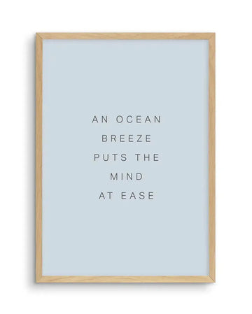 Framed art print with black sans-serif text on a pale blue background, displayed in a natural oak frame with white border. The text reads: "AN OCEAN BREEZE PUTS THE MIND AT EASE".