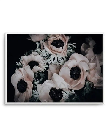 Framed art print featuring a close-up photograph of pale blush pink anemone flowers with dramatic black centers against a dark background. Soft green foliage and stems are visible between the blooms. The composition is intimate and moody, with shallow depth of field creating soft focus in the background. The print is displayed in a white frame with white border.